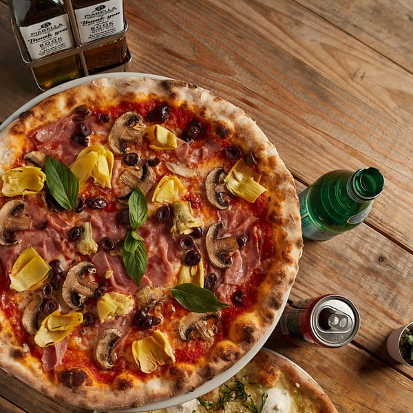 Piabella wood-fired pizza with ham, mushrooms, olives, artichokes, and basil on a wooden table.