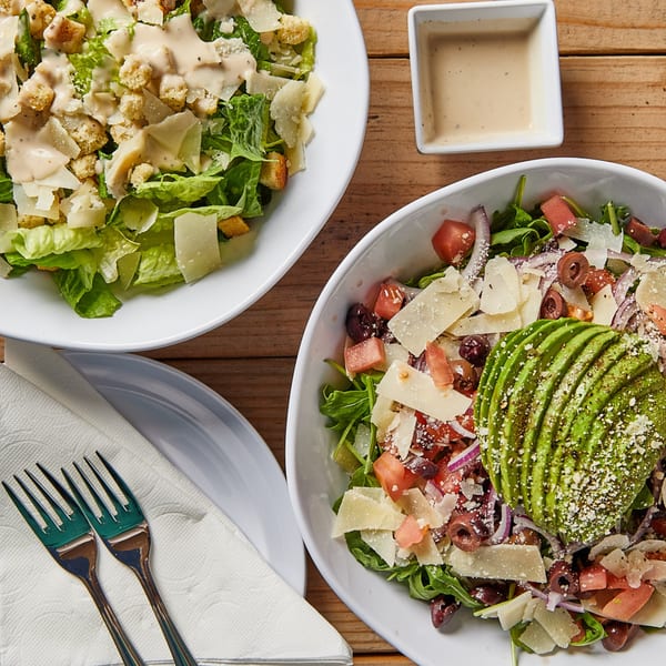 Two fresh salads, one Caesar and one with avocado, tomatoes, and olives, on a wooden table.
