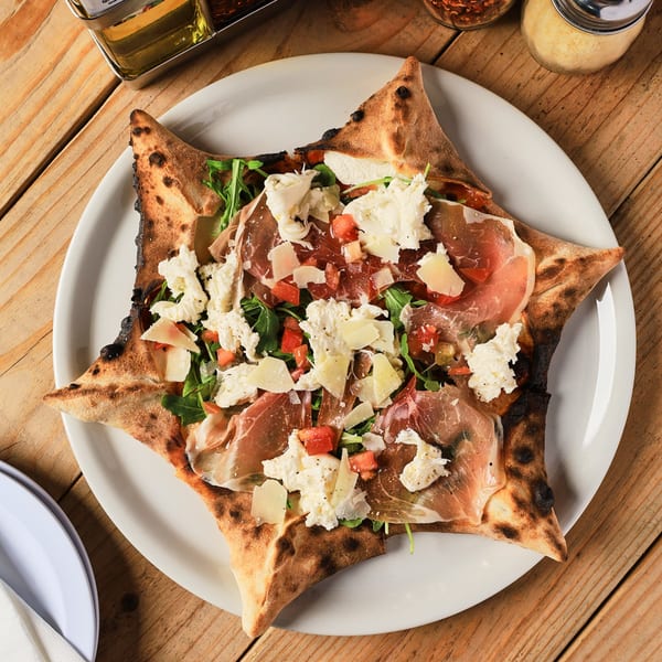 Star-shaped Piabella wood-fired pizza with prosciutto, burrata, arugula, and tomatoes on a wooden table.