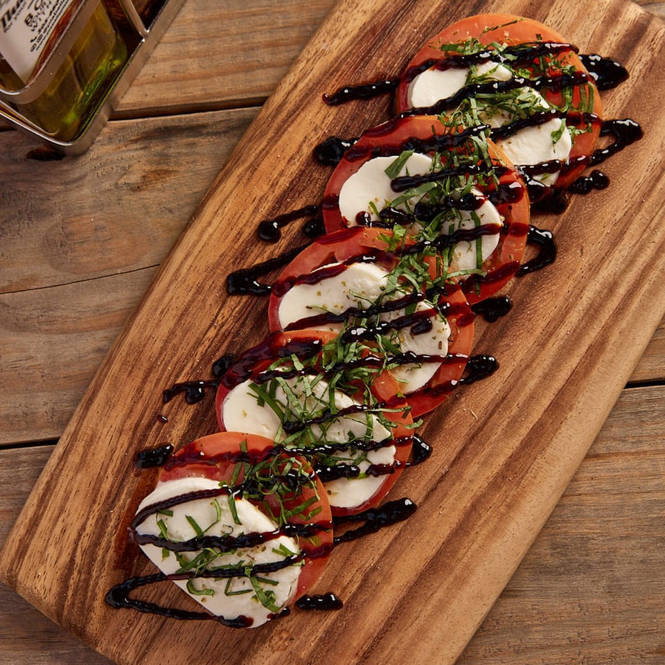 Caprese salad with sliced tomatoes, mozzarella, basil, and balsamic glaze on a wooden board.